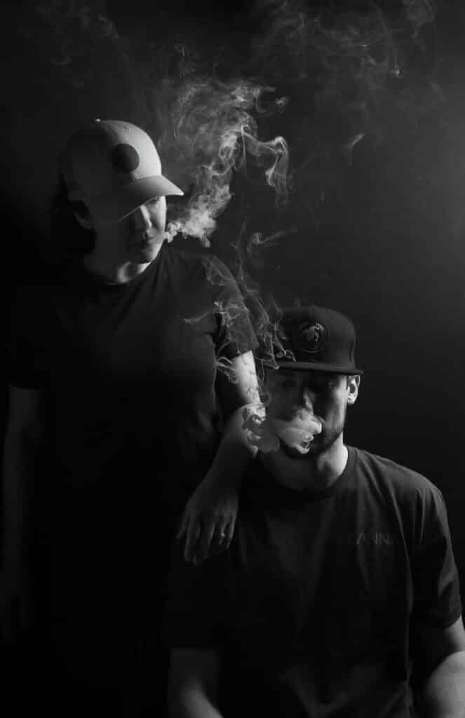 A person stands with an arm resting on the shoulder of another seated person, who is exhaling smoke—possibly from about kanna collective cbd. Both wear caps and dark clothing, with smoke swirling in the air.