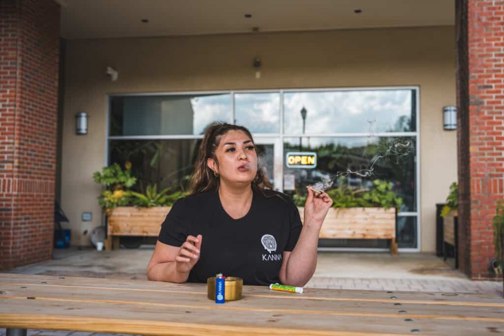 A person sits at an outdoor wooden table, smoking premium CBD, with a grinder and rolling papers in front of them. Plants and an "OPEN" sign are visible in the background.