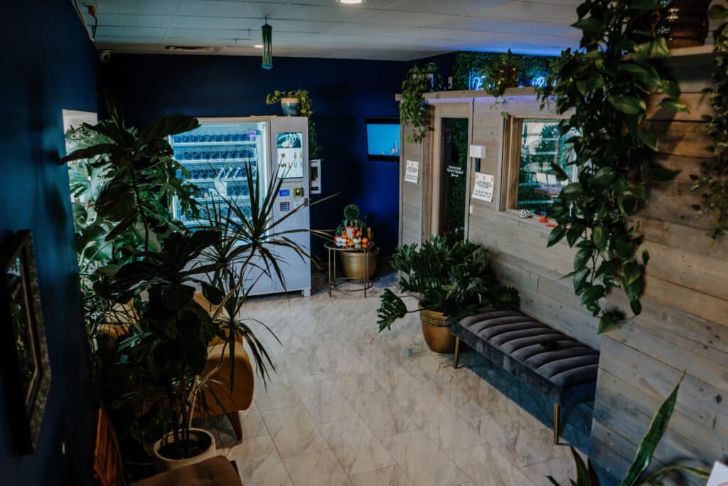 A waiting area with many green plants, a bench, and a vending machine against a blue wall, with bright lighting and tiled flooring.