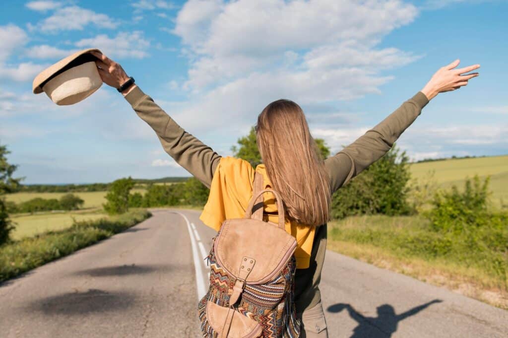 Person with long hair and a backpack stands on a rural road with arms raised, holding a hat, facing away from the camera toward green fields and a blue sky.