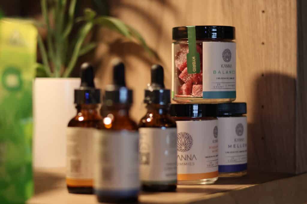 Several bottles and jars of wellness products, including dropper bottles and a jar of gummies, are displayed on a wooden shelf next to a potted plant.