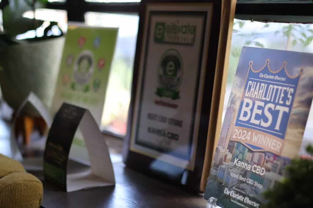 A display of framed awards and certificates on a windowsill, including a "Charlotte's Best 2024 Winner" plaque for Kanna CBD.