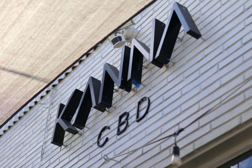A sign reading "KIMA CBD" in black letters is mounted on a white brick wall beneath an outdoor shade cloth.