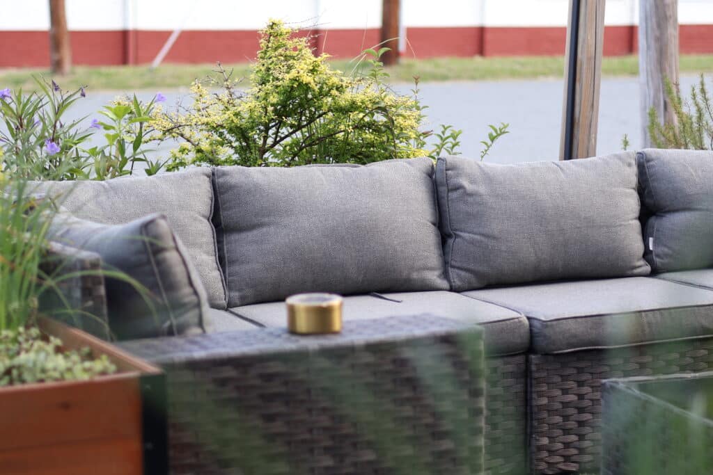 Gray outdoor sectional sofa with cushions next to a dark wicker table holding a small round tin, surrounded by green plants and a blurred background.