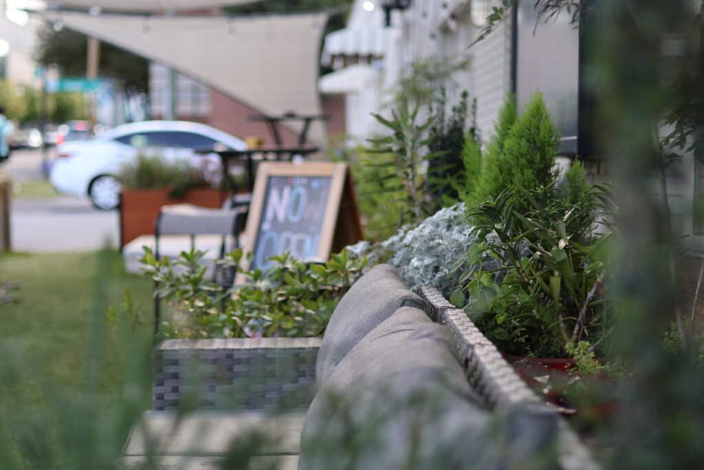 Outdoor seating area with a cushioned bench, green plants, and a wooden sign displaying "NO OPEN" near a street with parked cars.